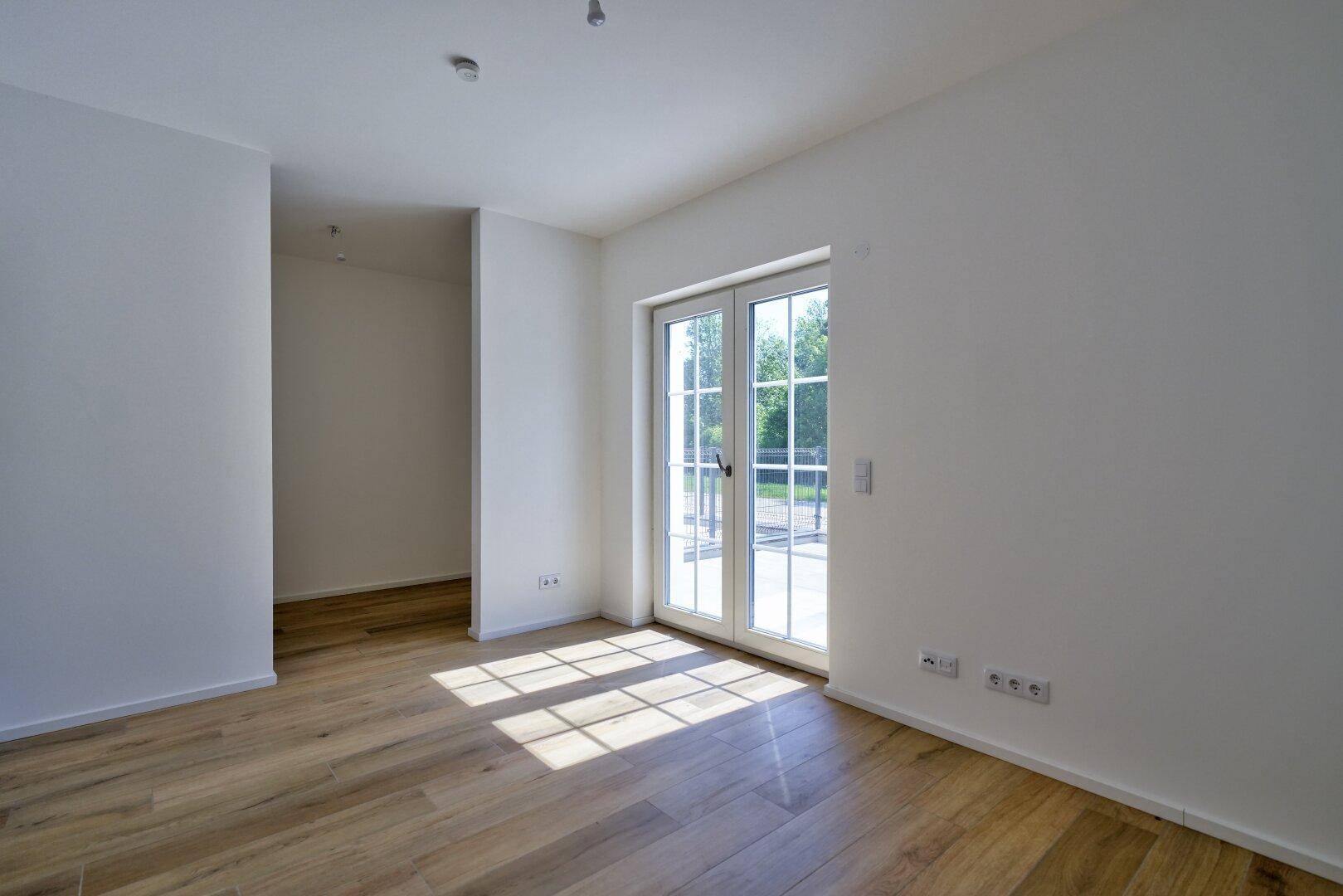 Lichtdurchfluteter Raum mit Holzboden und großen Fenstertüren, die Sonnenlicht hereinlassen.