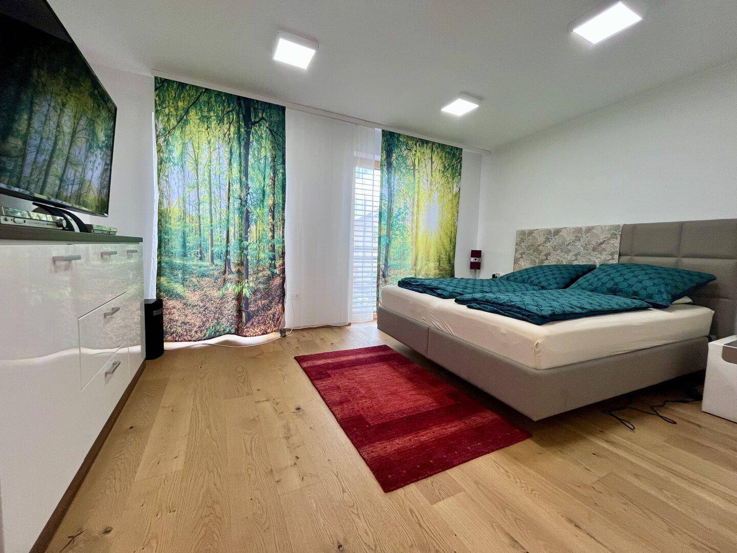 Schlafzimmer mit Holzboden, großem Bett und dekorativen Vorhängen mit Waldmotiv.