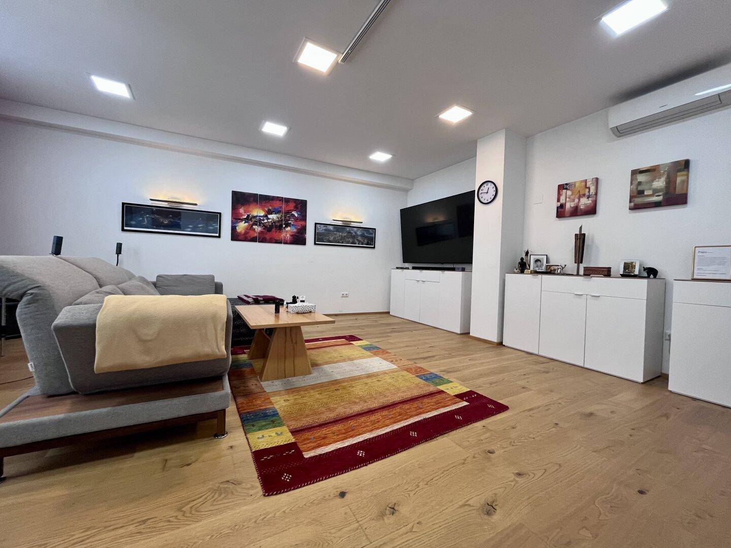 Geräumiges Wohnzimmer mit großem Fernseher, bequemer Couch und farbenfrohem Teppich auf Holzparkett.