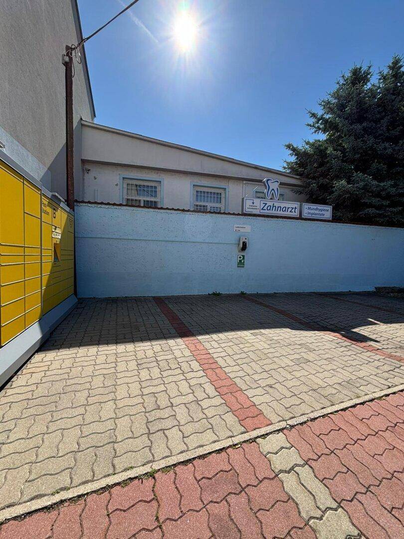 Blick auf den Parkplatz mit einer Ladestation, einem Zahnarzt-Schild und einer Paketstation unter blauem Himmel.