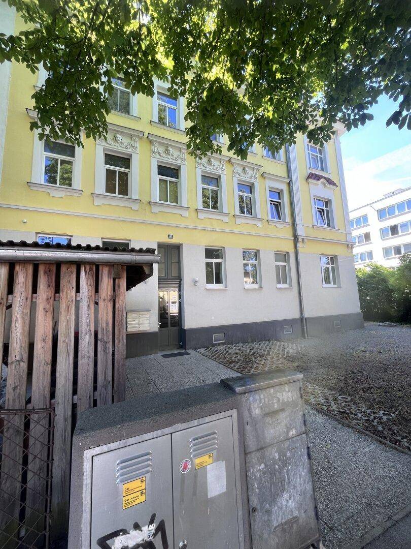 Gebäudefassade mit Eingangsbereich und Versorgungsboxen im Vordergrund, umgeben von Bäumen.