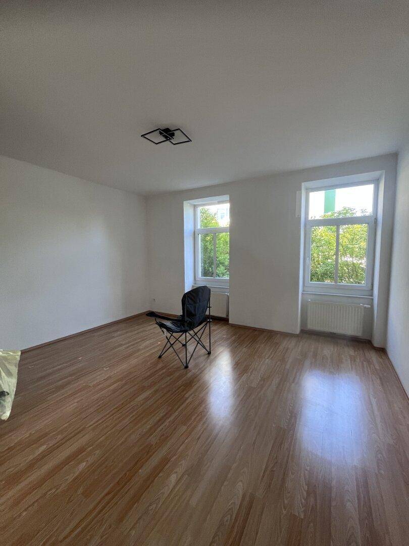 Leerer Raum mit Laminatboden, weißen Wänden und zwei großen Fenstern mit Blick ins Grüne.