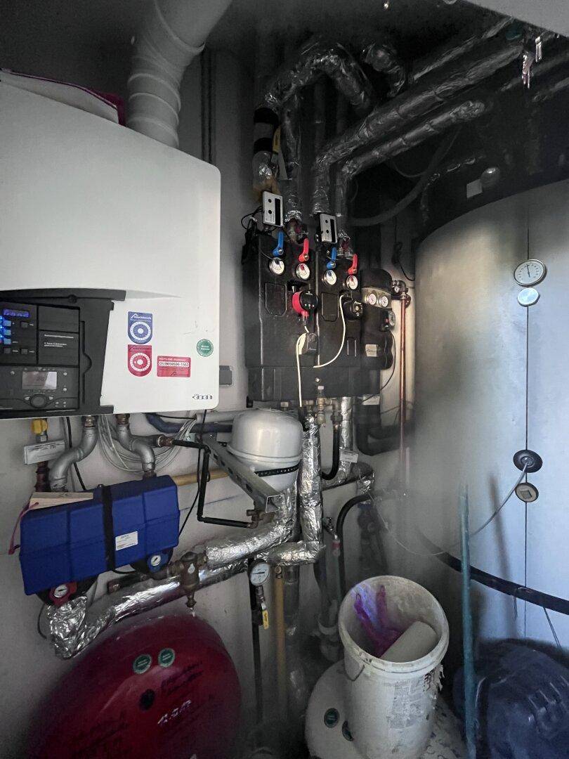 Heizungsraum mit komplexen Rohrleitungen, einem Warmwasserspeicher und verschiedenen Geräten.
