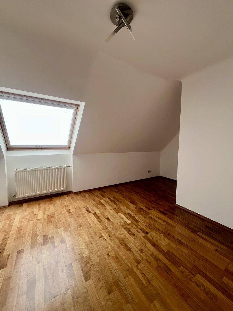 Leeres Zimmer im Dachgeschoss mit Holzboden, großem Dachfenster und Heizkörper.