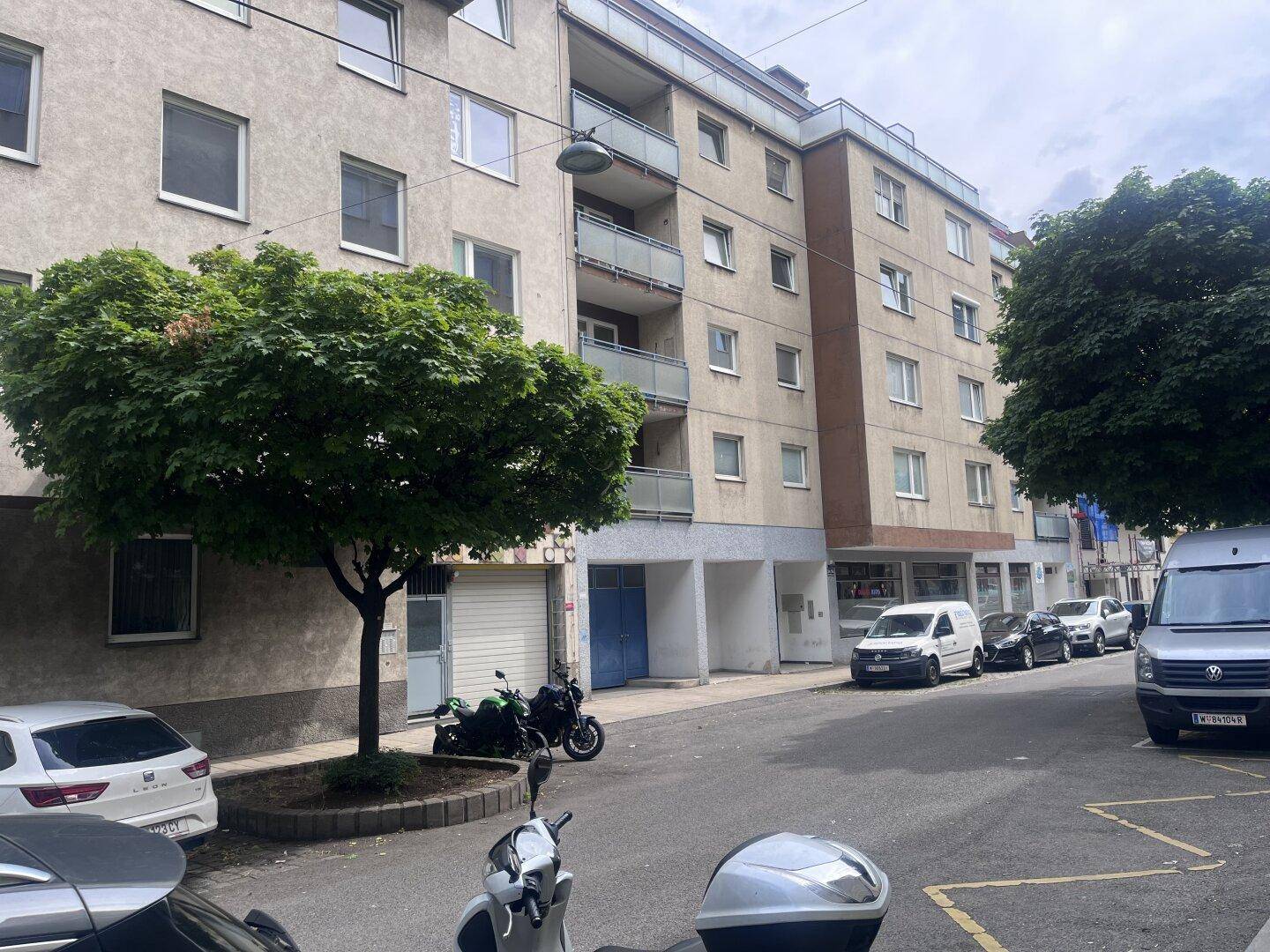 Mehrfamilienhaus in städtischer Umgebung mit Balkonen und geparkten Fahrzeugen auf der Straße.