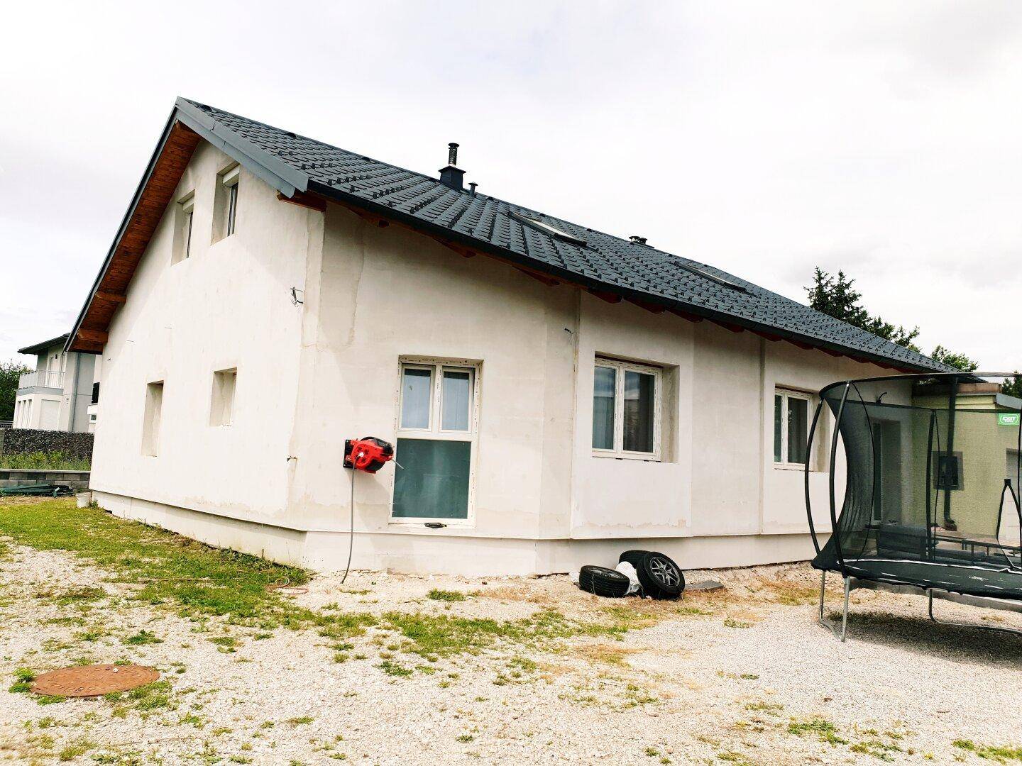 Seitenansicht des Hauses mit weißer Fassade, dunklem Dach und einem Trampolin im Garten.