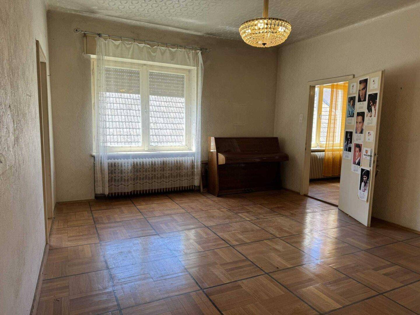 Wohnzimmer mit Parkettboden, Fenster mit Jalousien, einem Klavier und Zugang zu einem weiteren Raum.