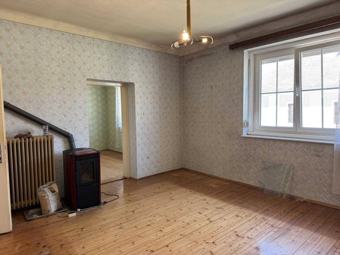 Wohnraum mit Holzdielenboden, gemusterter Tapete, einem Holzofen und einem Fenster.