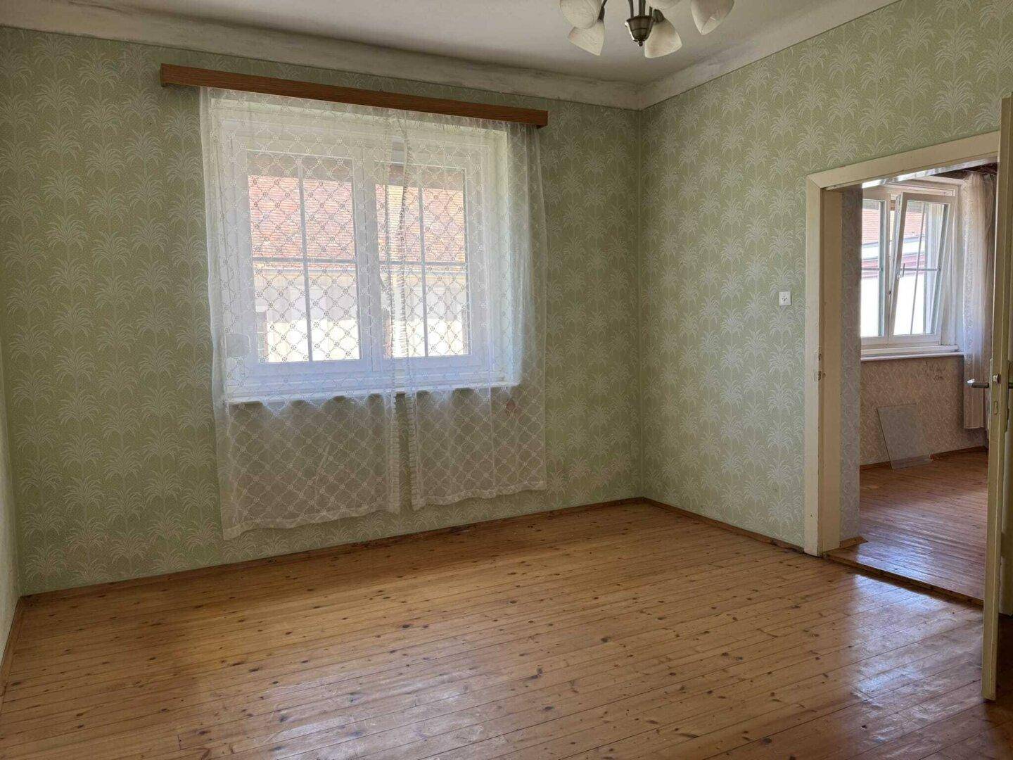 Schlafzimmer mit Holzdielenboden, grüner gemusterter Tapete und einem Fenster mit Vorhang.