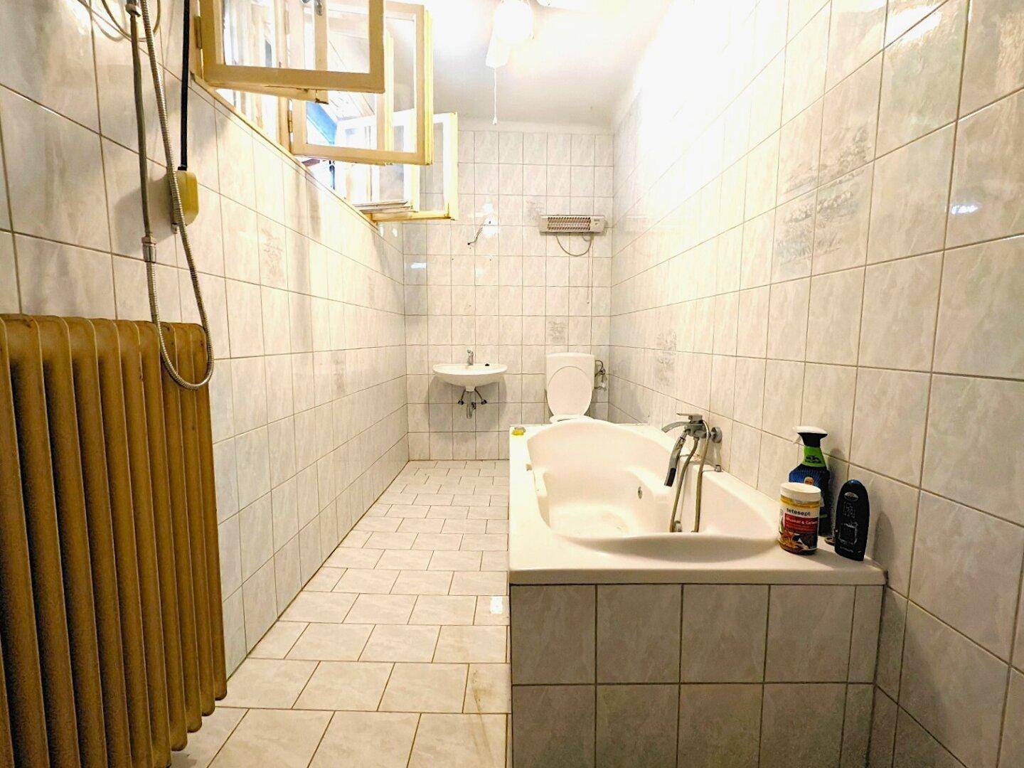 Badezimmer mit weißen Fliesen, Badewanne, Waschbecken, Toilette und einem Fenster.