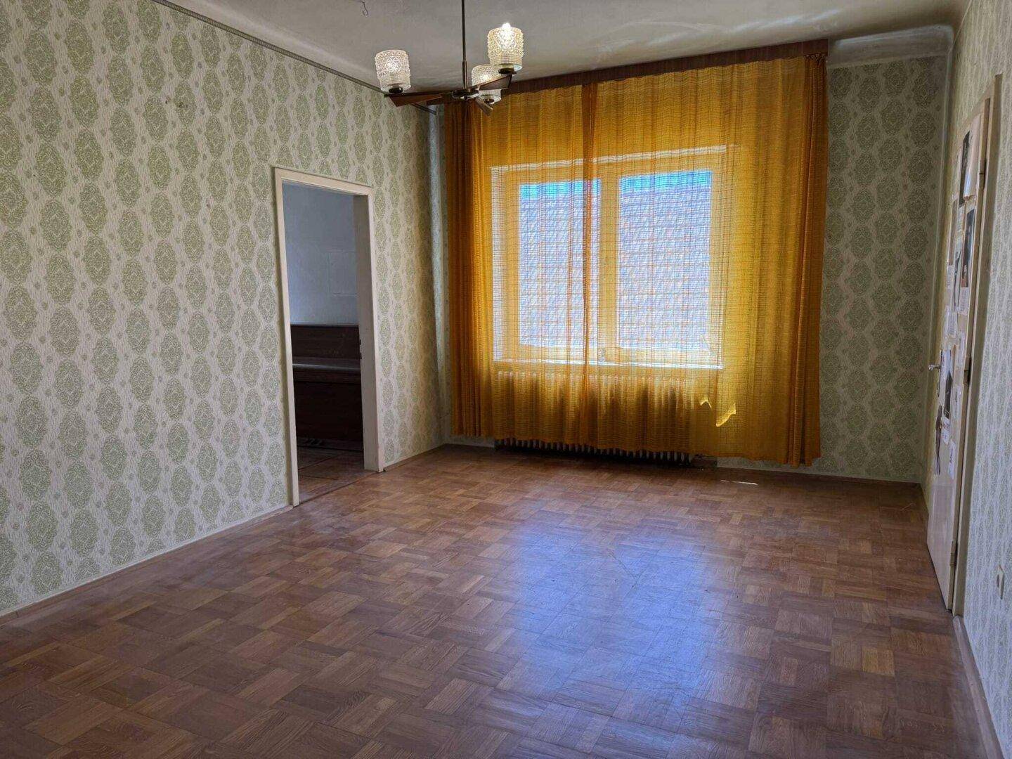Wohnzimmer mit Parkettboden, gemusterter Tapete und einem Fenster mit gelben Vorhängen.