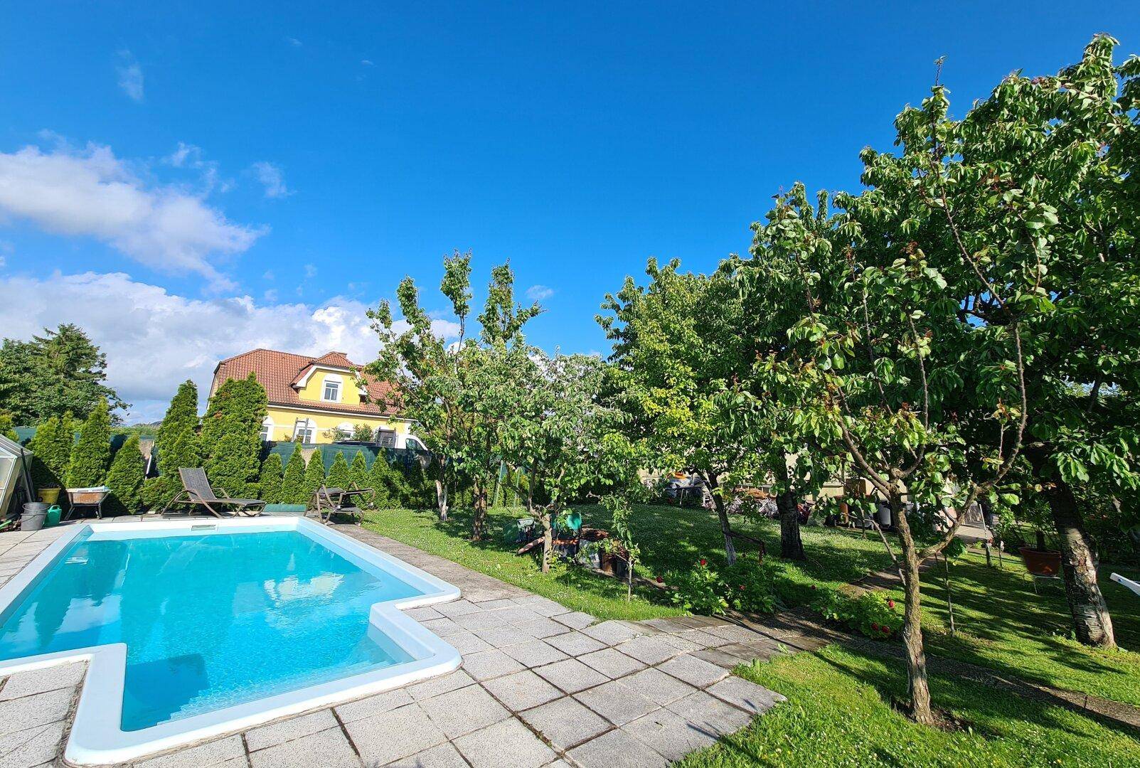 Garten mit Pool und Liegeflächen, umgeben von Bäumen und einem Haus im Hintergrund.