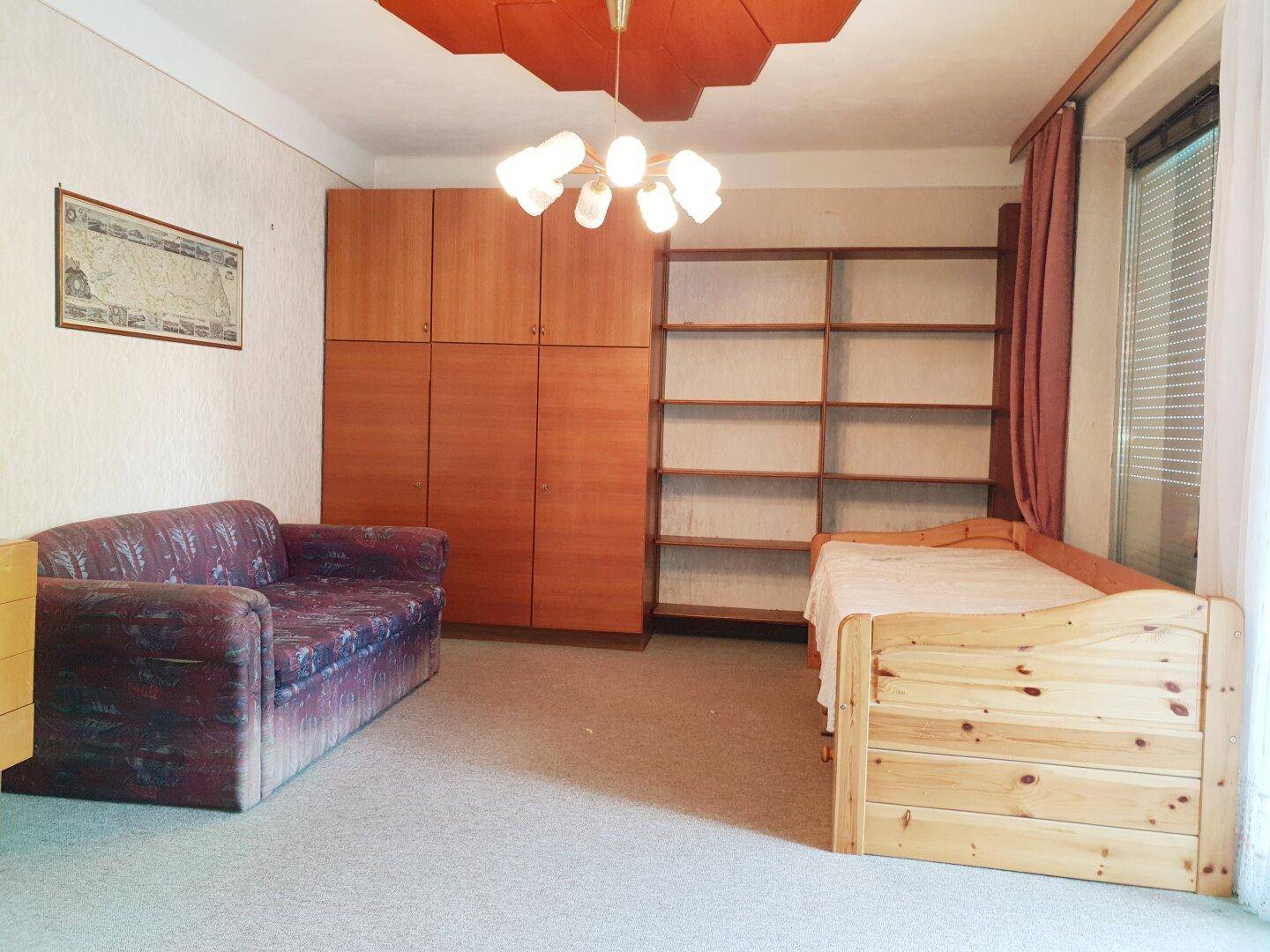 Schlafzimmer mit Einzelbett, Kleiderschrank und Bücherregal, Teil eines größeren Raumes.