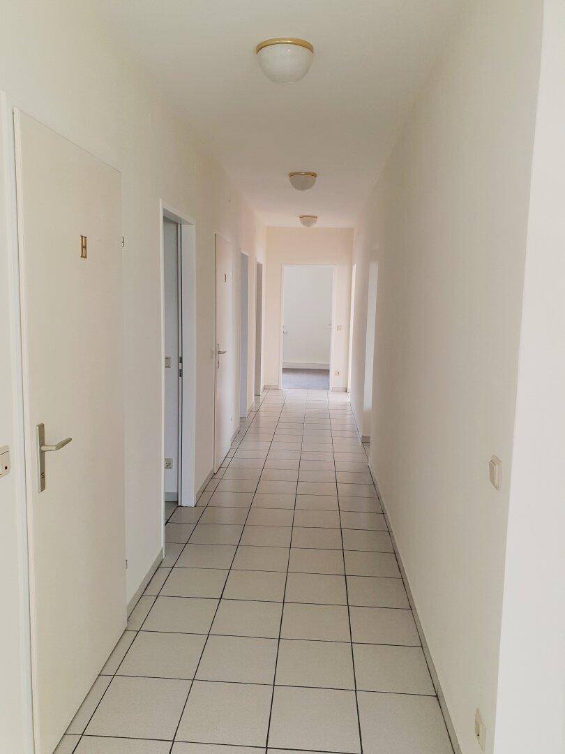 Langer, heller Flur mit weißen Fliesen, mehreren Türen und Deckenleuchten, führt durch die Wohnung.