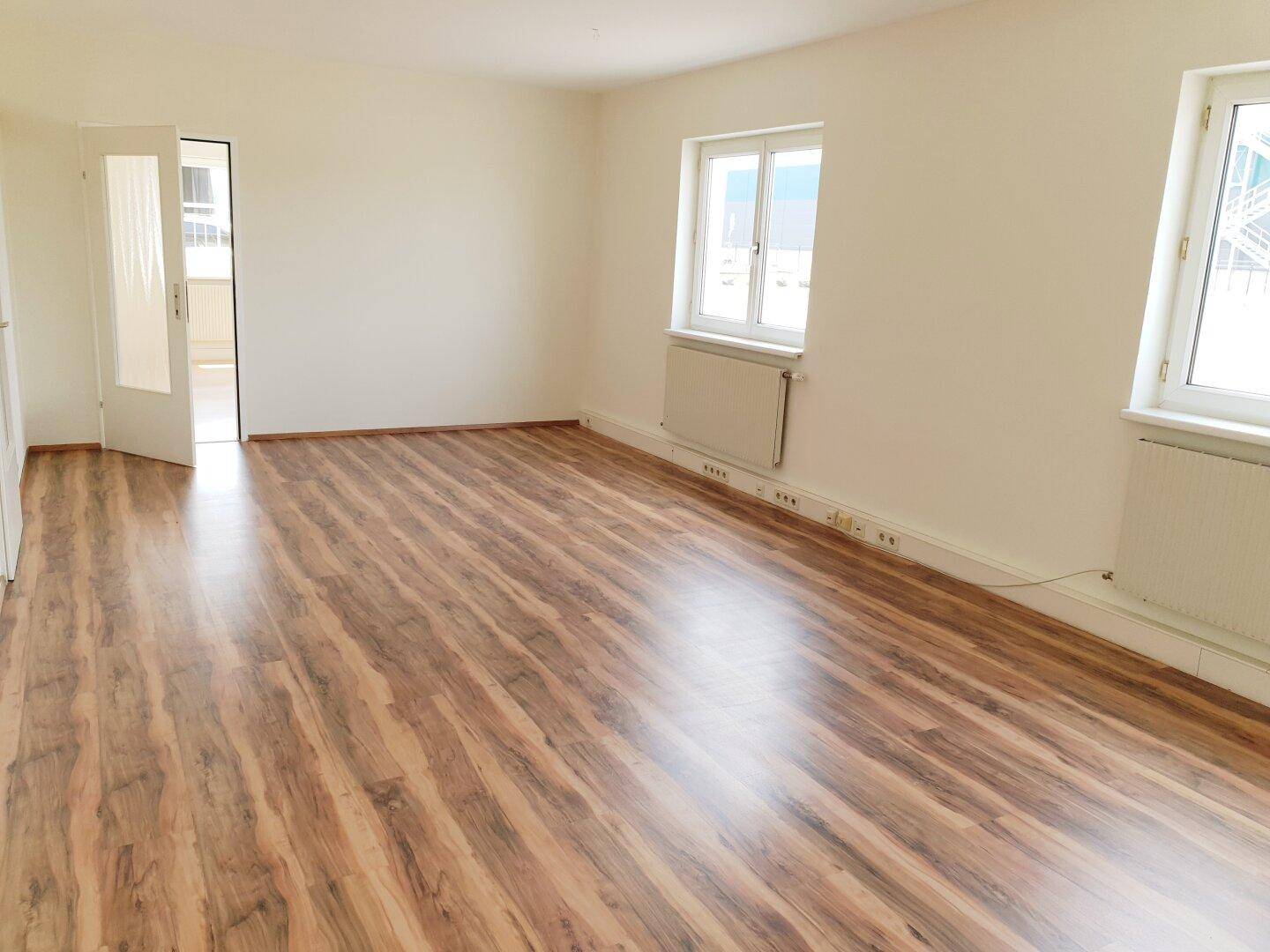 Heller, leerer Raum mit Laminatboden, weißen Wänden und zwei Fenstern, ideal als Wohnzimmer.