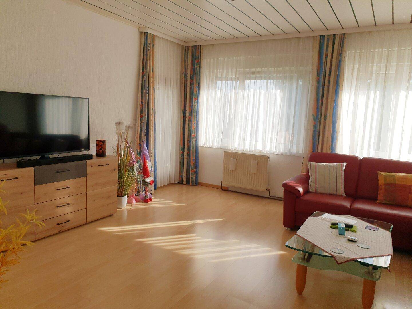 Gemütliches Wohnzimmer mit hellem Laminatboden, rotem Sofa und viel Tageslicht.
