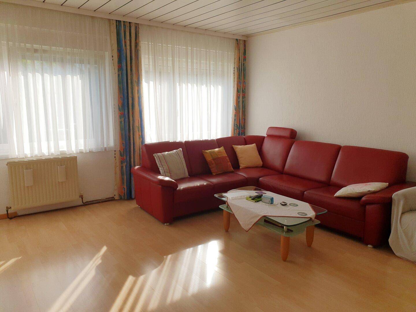 Geräumiges Wohnzimmer mit rotem Ecksofa, hellem Laminatboden und großen Fenstern.
