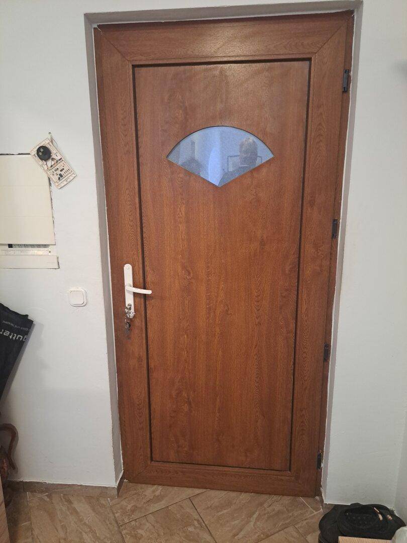 Braune Holztür mit einem kleinen, fächerförmigen Glaseinsatz und weißem Griff.