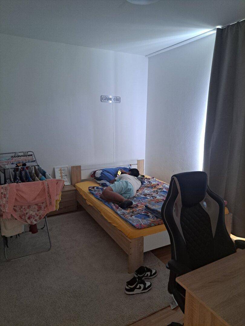 Kinderzimmer mit Einzelbett, Schreibtisch und Teppichboden in hellem Grau.