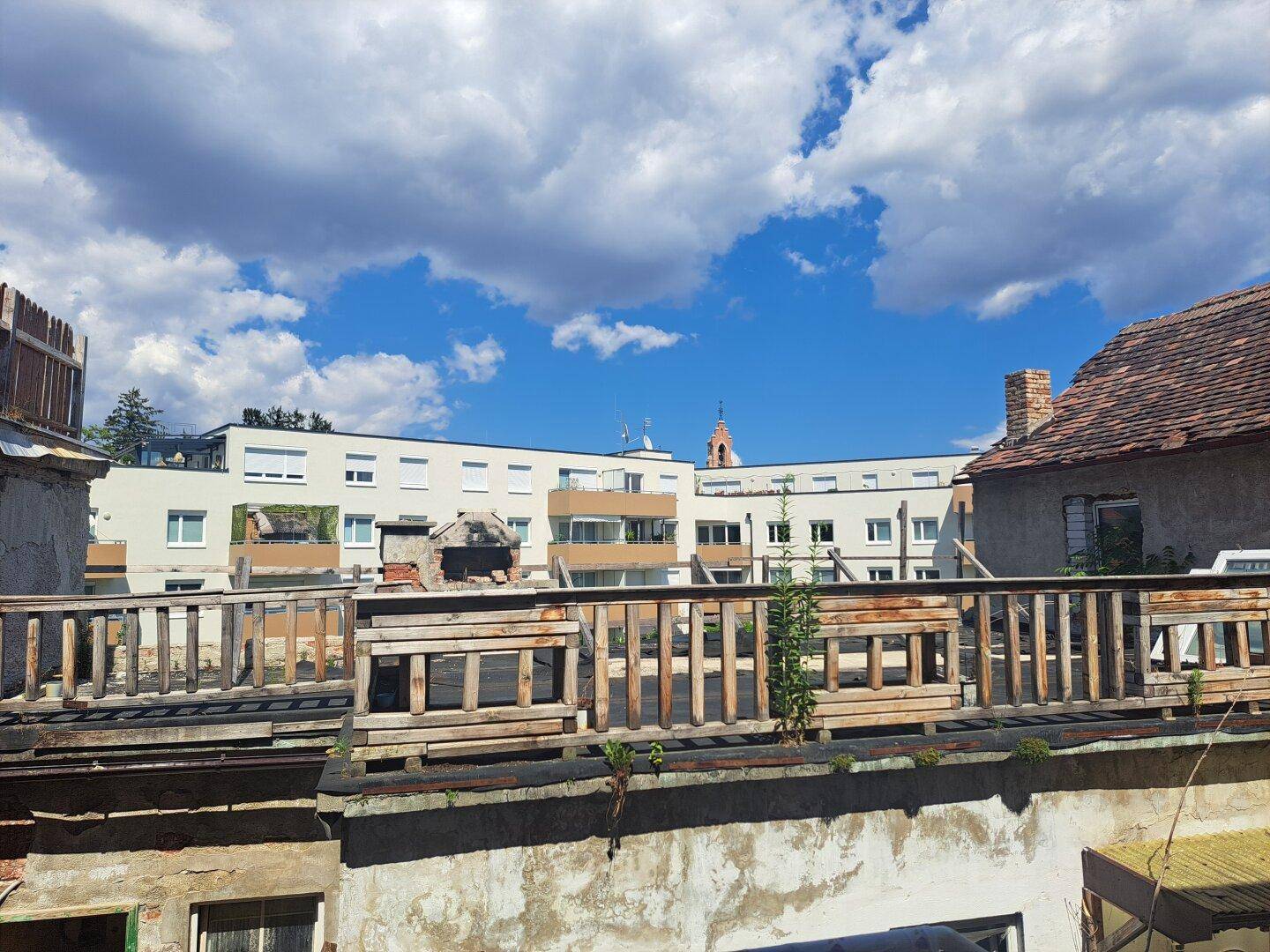Blick über alte Dächer auf moderne Wohngebäude und einen Kirchturm im Hintergrund.
