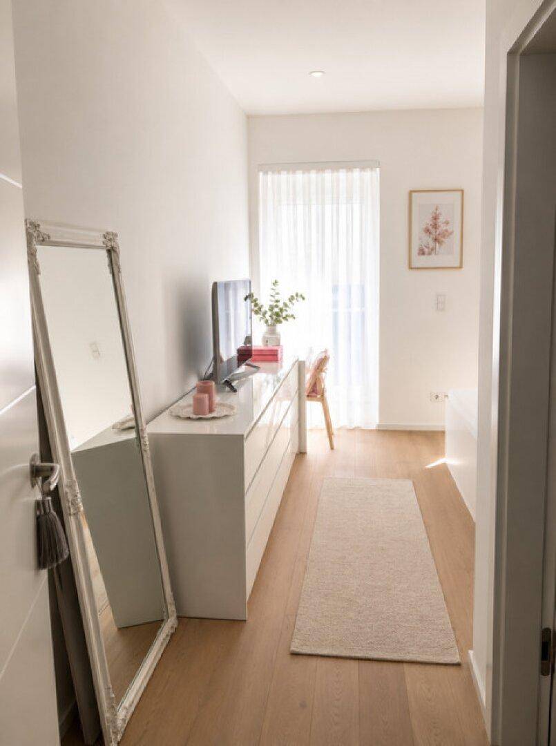 Heller Flur mit Holzboden, großem Spiegel und Kommode, der zu einem Fenster führt.