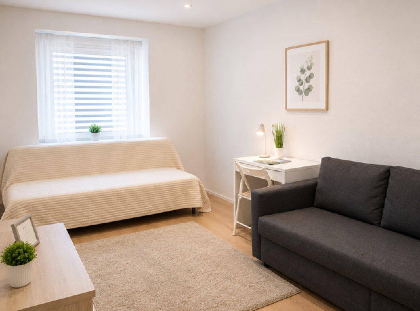 Helles Wohnzimmer mit beigem Sofa, Schreibtisch und großem Fenster für viel Tageslicht.