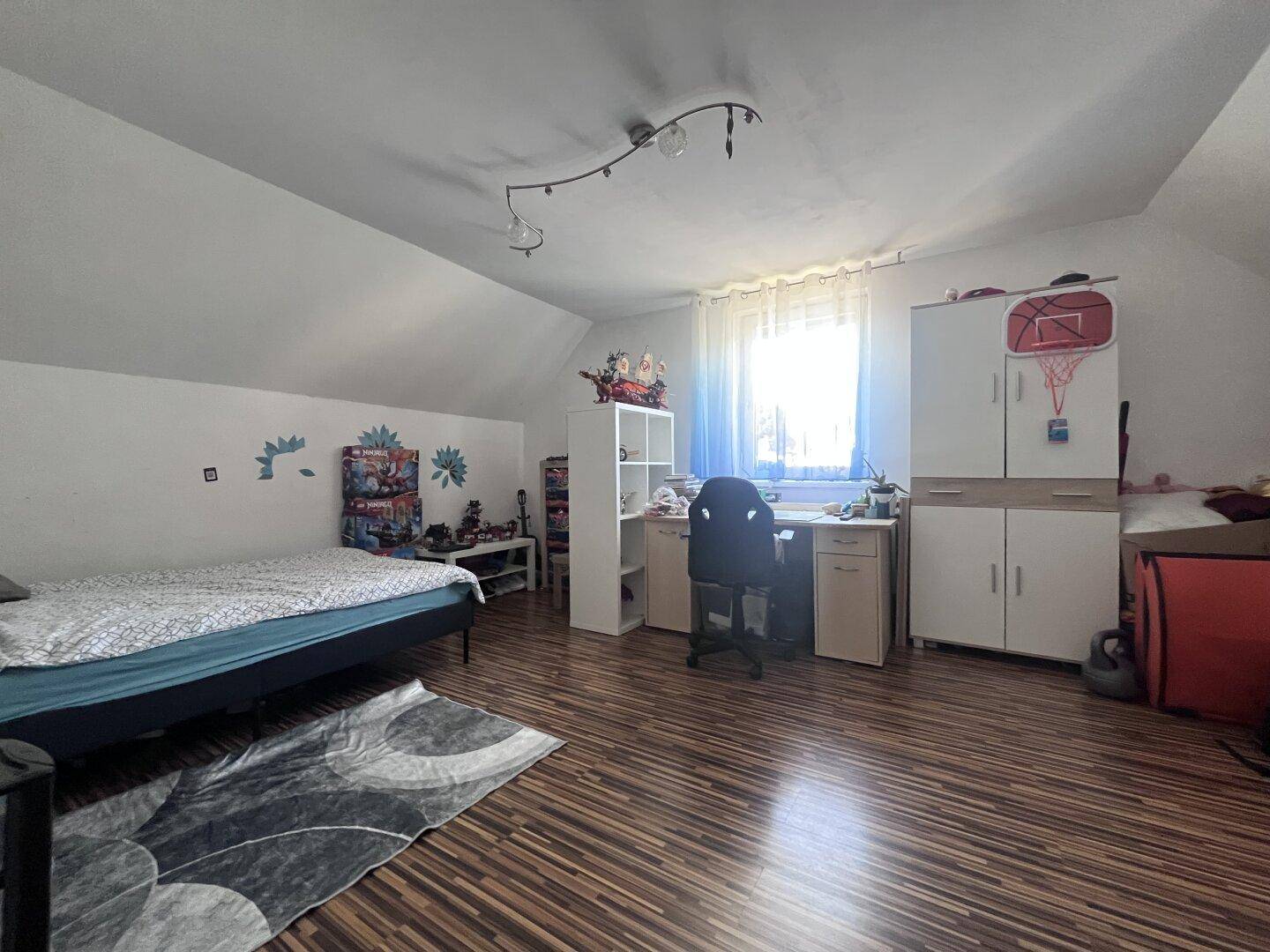 Helles Kinderzimmer im Dachgeschoss mit Schreibtisch, Bett und Laminatboden, gut beleuchtet.