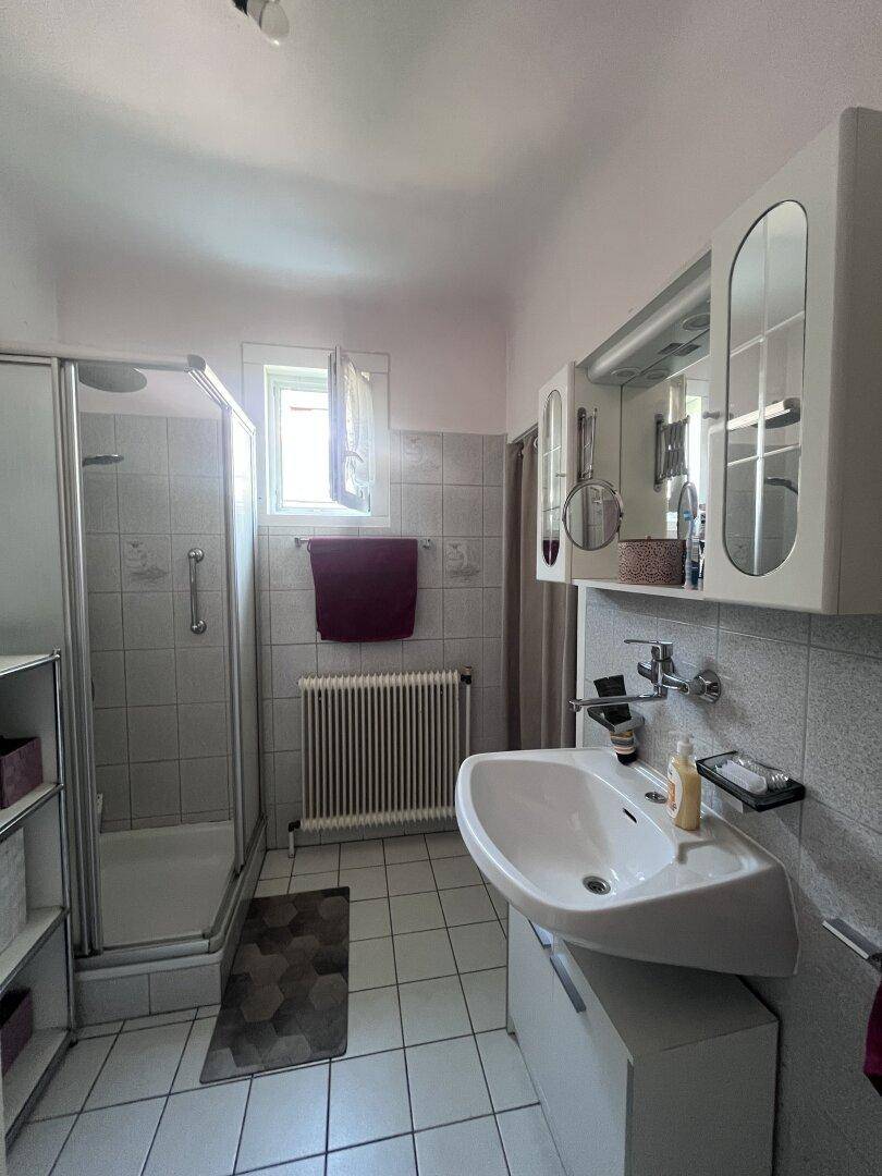 Badezimmer mit Dusche, Waschbecken und Fenster, ausgestattet mit hellen Fliesen.