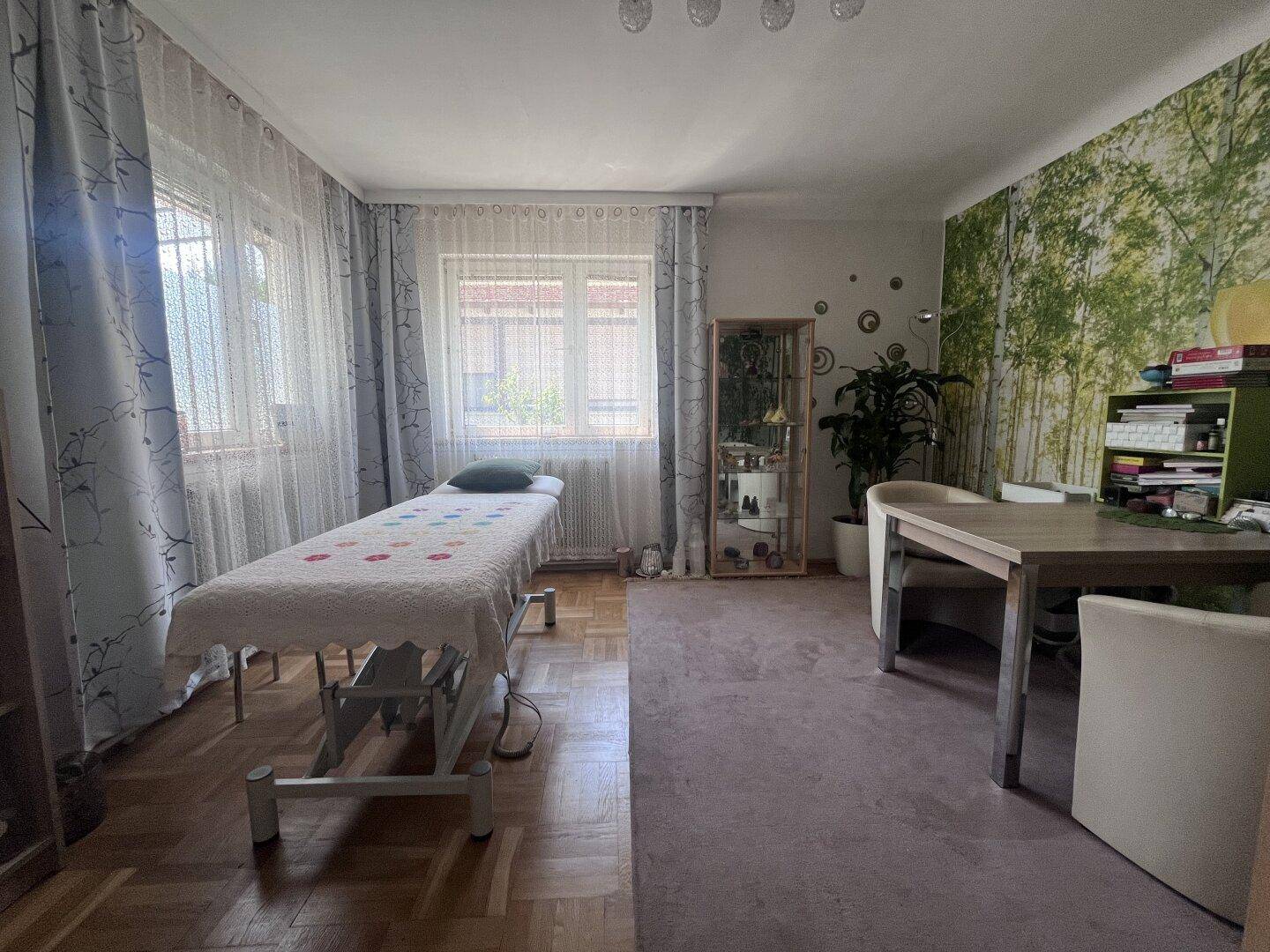 Heller Raum mit großen Fenstern, Waldtapete und einer Behandlungsliege, vielseitig nutzbar.
