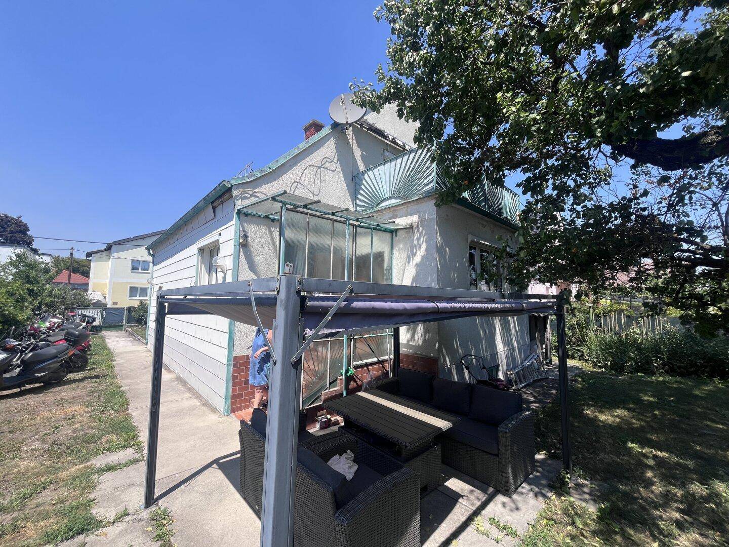 Außenbereich mit überdachter Terrasse und Gartenmöbeln, umgeben von Grün und Bäumen.