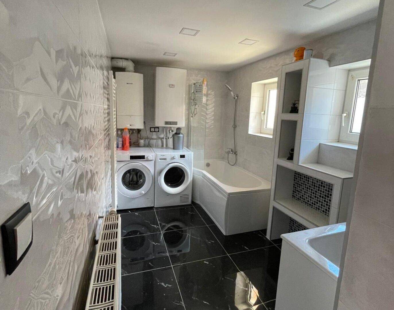 Modernes Badezimmer mit Badewanne, Waschmaschine, Trockner und schwarzen Fliesen.
