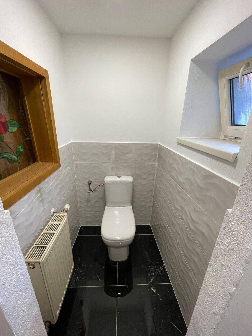 Separate Toilette mit weißen Wänden, schwarzen Bodenfliesen und kleinem Fenster.