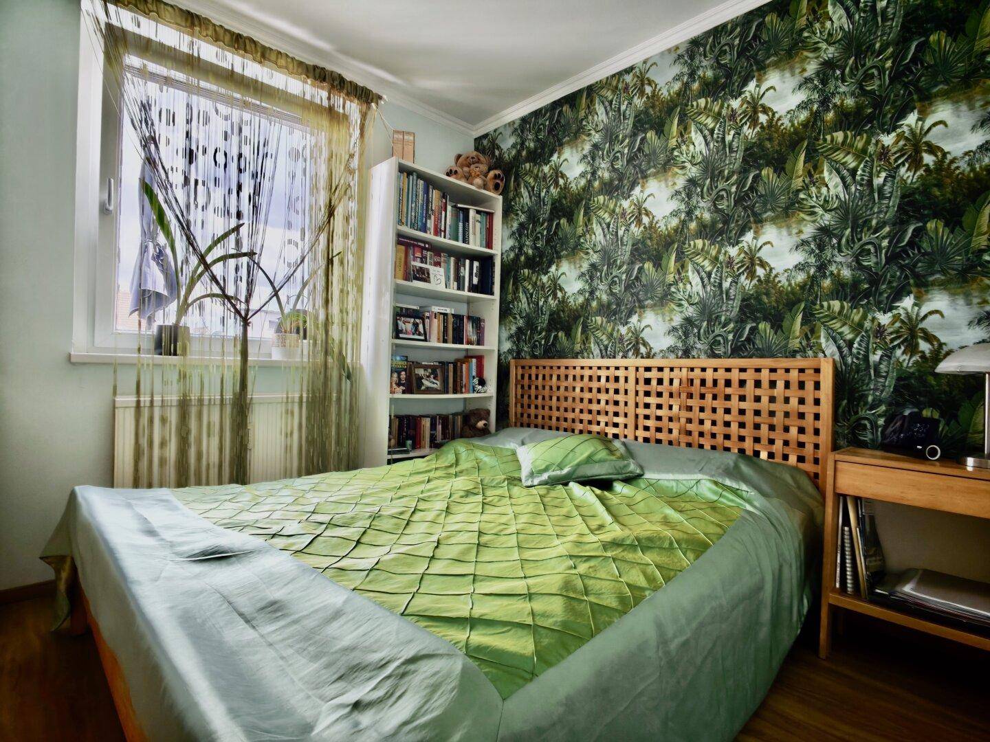 Schlafzimmer mit Holzbett, Bücherregal und auffälliger Tapete mit Dschungelmotiv.
