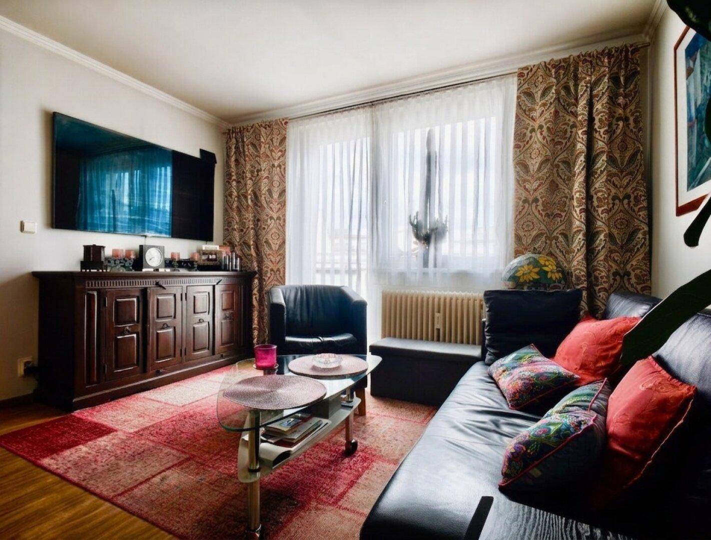Helles Wohnzimmer mit schwarzem Ledersofa, Couchtisch und Fenster mit gemusterten Vorhängen.