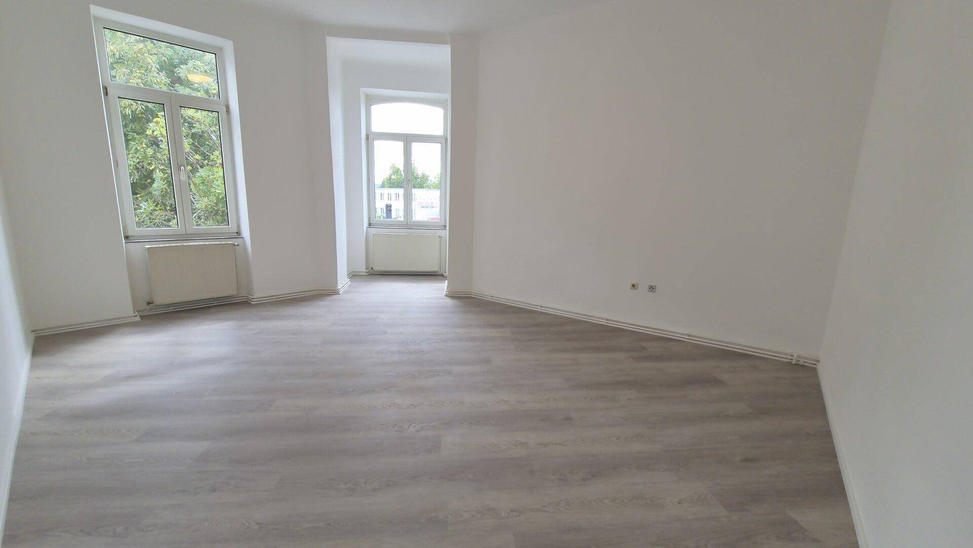 Helles, leeres Zimmer mit modernem Laminatboden und zwei Fenstern mit Blick ins Grüne.