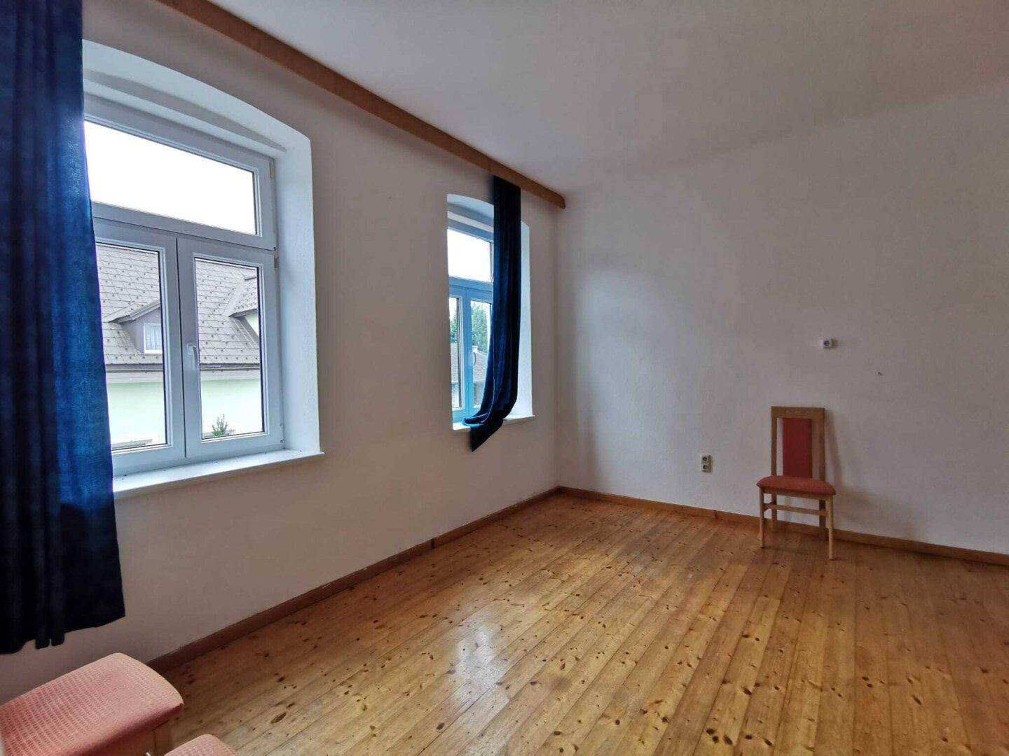 Schlichter Raum mit Holzdielenboden, weißen Wänden und zwei Fenstern mit blauen Vorhängen.