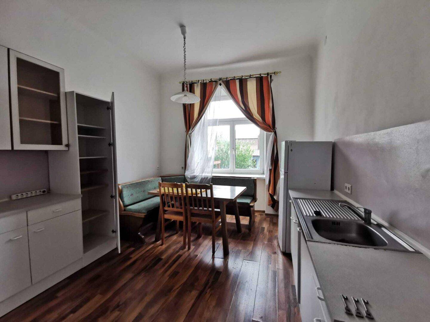 Wohnküche mit weißen Schränken, Essecke, Fenster mit gestreiften Vorhängen und Kühlschrank.
