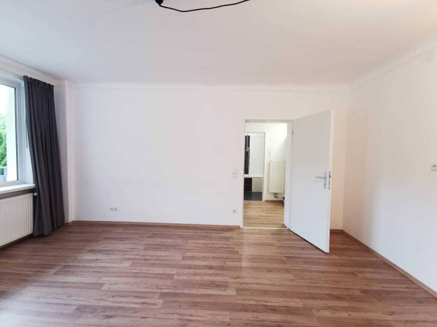 Helles Zimmer mit Laminatboden, großem Fenster und Zugang zum Badezimmer.
