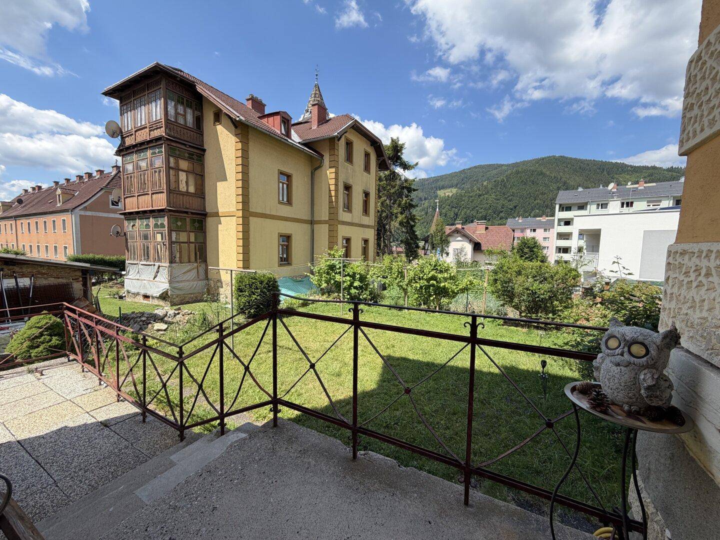 Blick vom Eingangsbereich auf den Garten und das historische Gebäude mit Bergblick im Hintergrund.