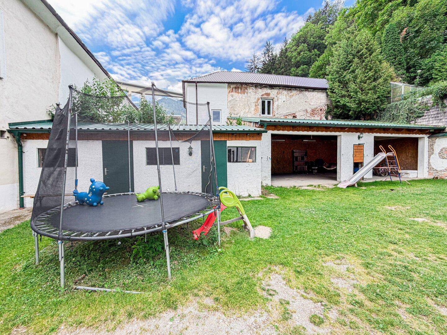 Garten mit Trampolin, Rutsche und Rasenfläche, umgeben von Natur.