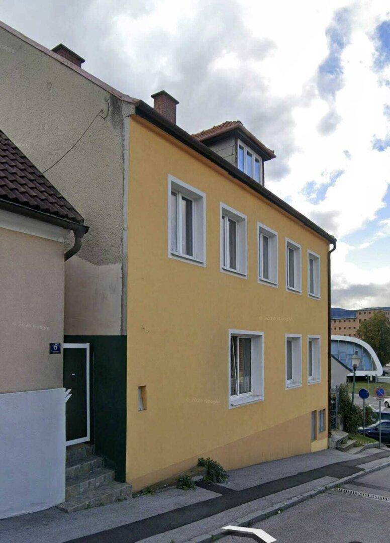Ansicht der Gebäudeseite mit einem dunklen Eingang und weiteren Fenstern unter bewölktem Himmel.
