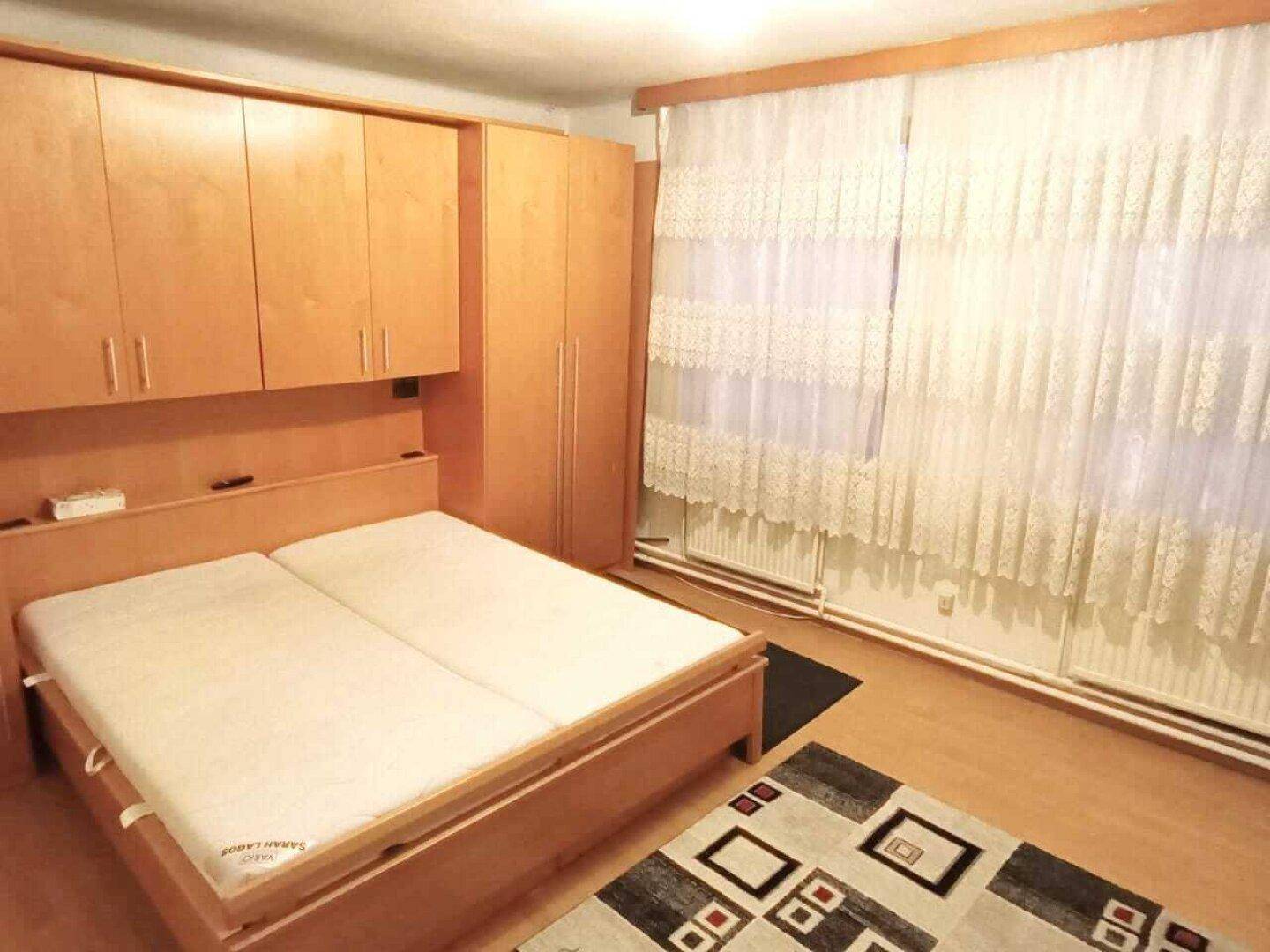 Schlafzimmer mit Doppelbett, Einbauschränken aus Holz und großen Fenstern mit Vorhängen.
