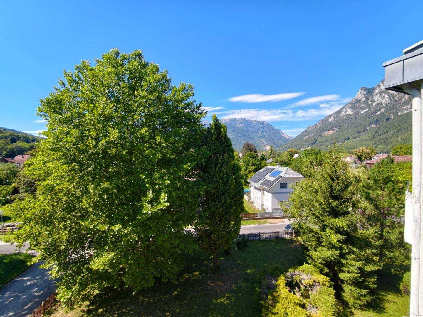 Weitläufiger Blick auf grüne Bäume, umliegende Häuser mit Solaranlagen und majestätische Berge im Hintergrund.