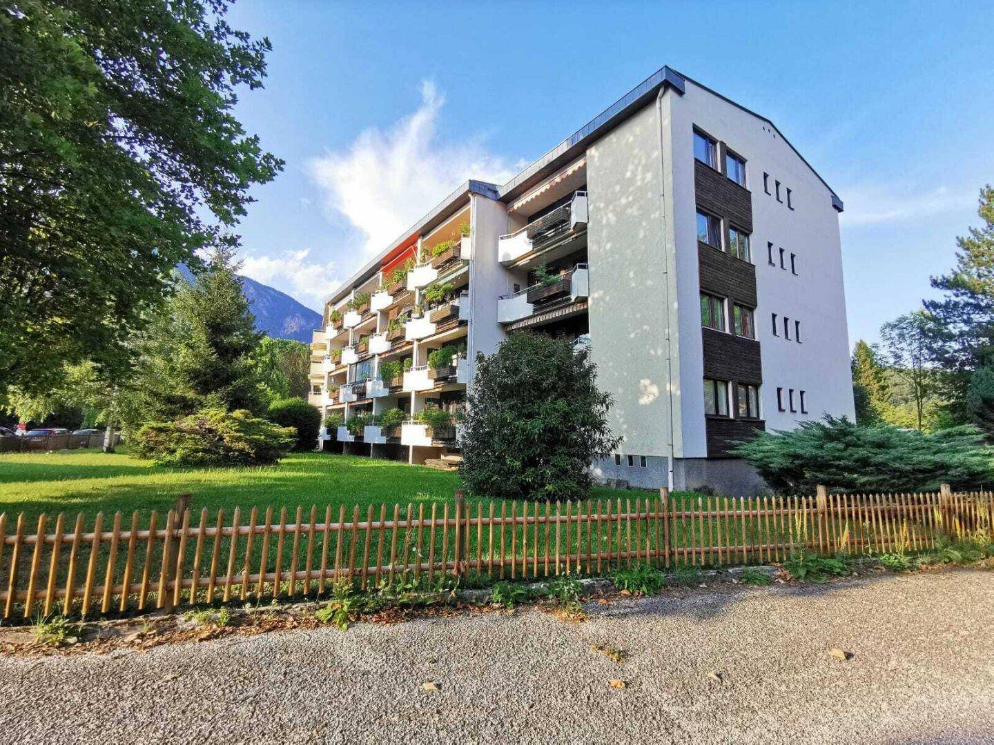 Mehrfamilienhaus mit vielen Balkonen und dunklen Holzverkleidungen, umgeben von einer grünen Wiese und Bäumen.
