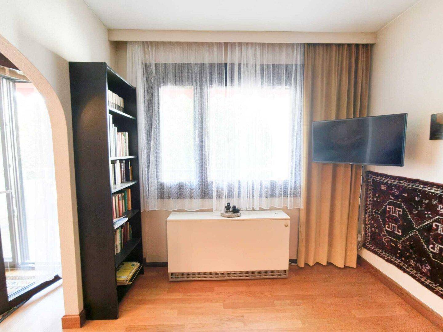 Helles Zimmer mit Holzfußboden, einem Bücherregal und einem Fernseher, durch ein Fenster gut beleuchtet.