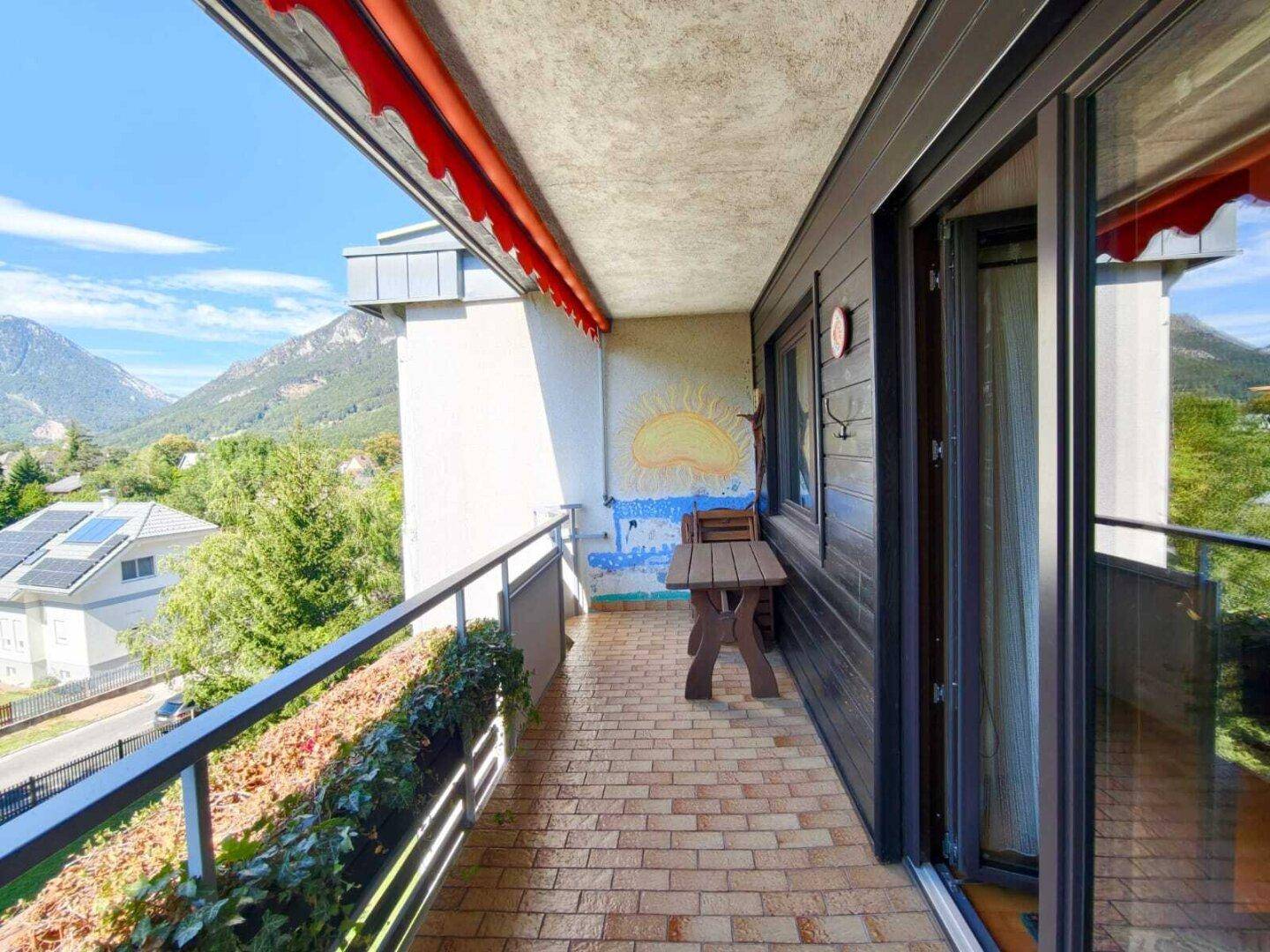 Langer Balkon mit Ziegelboden, einem Holztisch und Bergblick, geschützt durch eine rote Markise.