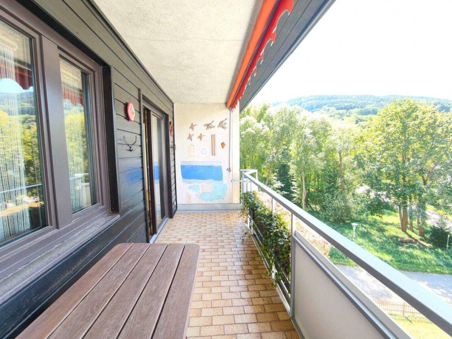 Geräumiger Balkon mit Ziegelboden, Holztisch und Blick auf grüne Hügel, geschützt durch eine Markise.
