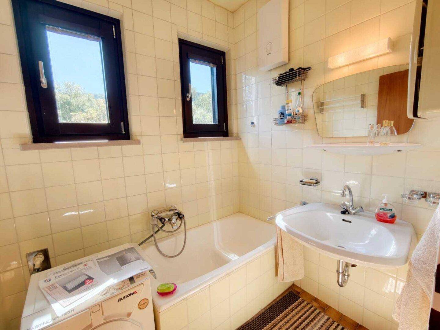 Helles Badezimmer mit Badewanne, Waschbecken und zwei Fenstern, ausgestattet mit hellen Fliesen.