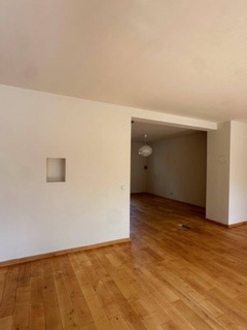 Großer, heller Raum mit Holzboden und offenem Übergang zu einem weiteren Bereich.