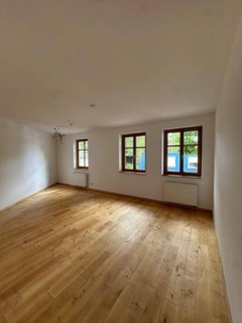 Helles Zimmer mit Holzboden und drei Fenstern, die viel Tageslicht hereinlassen und einen Blick ins Grüne bieten.